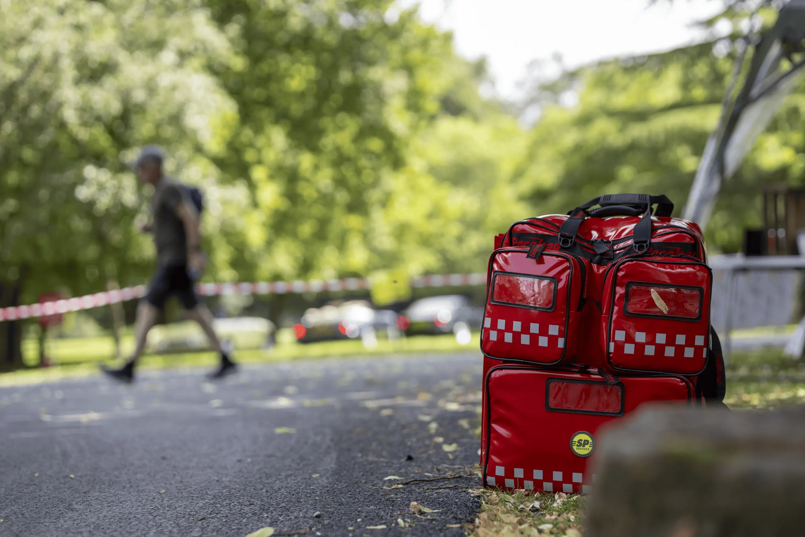 Red medical response bag positioned at an outdoor event site with a footpath and trees in the background.