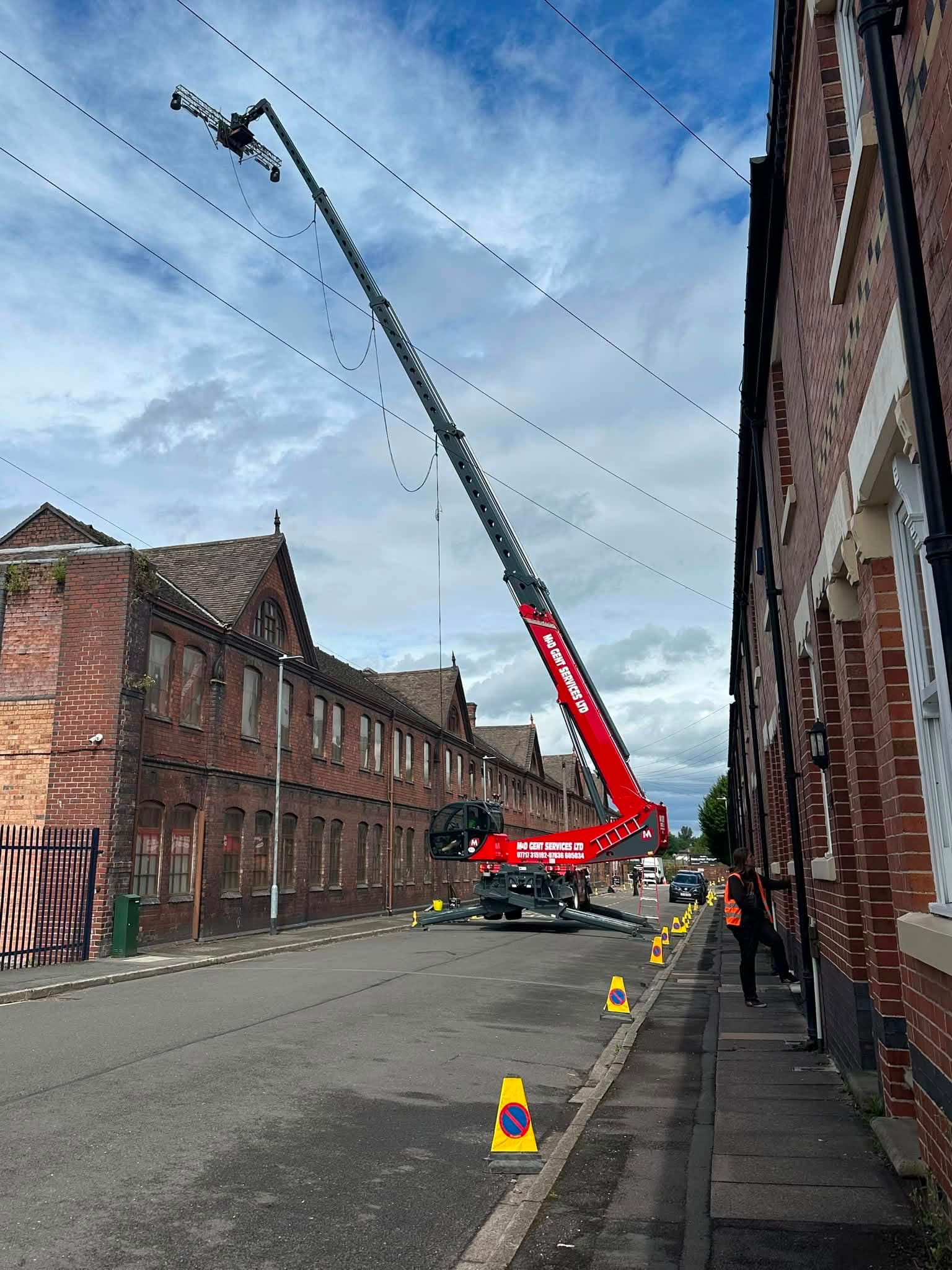 Film rigging cherry picker extended between brick buildings on a location street with safety cones and crew.