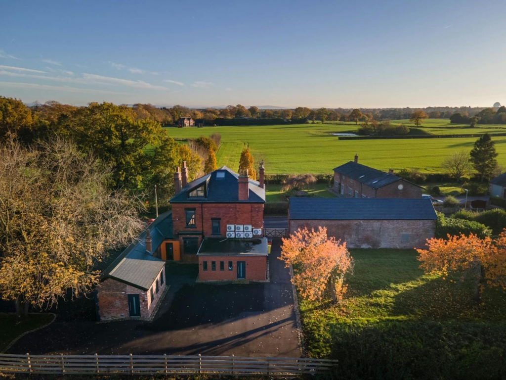 Aerial view of a two-story office building with red brick walls and a dark roof, situated in a rural area. The building is surrounded by lush green fields, with autumnal trees in vibrant shades of orange and yellow. A well-maintained driveway with a wooden fence leads to the property, which is bathed in the soft glow of the early morning or late afternoon sun."